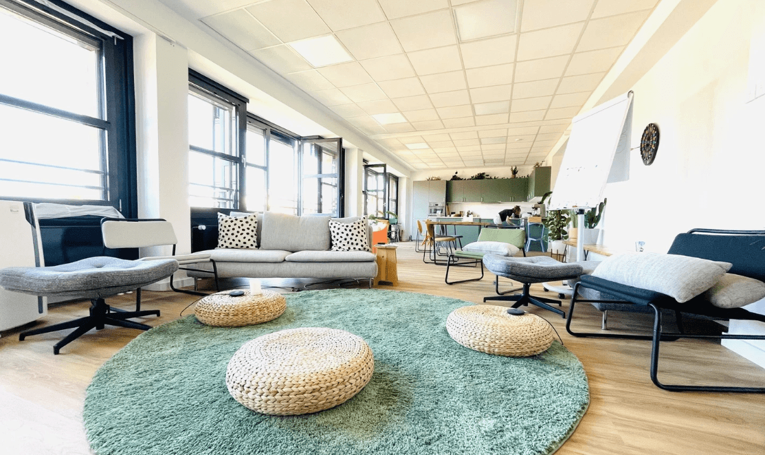 A bright and modern office lounge with cozy seating, a green rug, and an open kitchen area in the background.