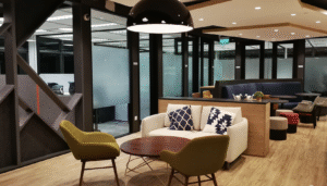 Cozy office lounge with wooden floor, white couch, green chairs, and glass-walled meeting rooms.